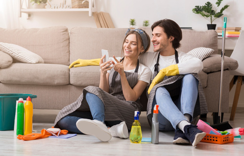 Couple sitting on floor smiling at phone surrounded by cleaning supplies